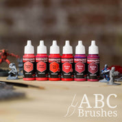 Army Painter Fanatics Cool Reds Triad on a wooden surface. There are wargaming models and an ABC paint brush next to them.