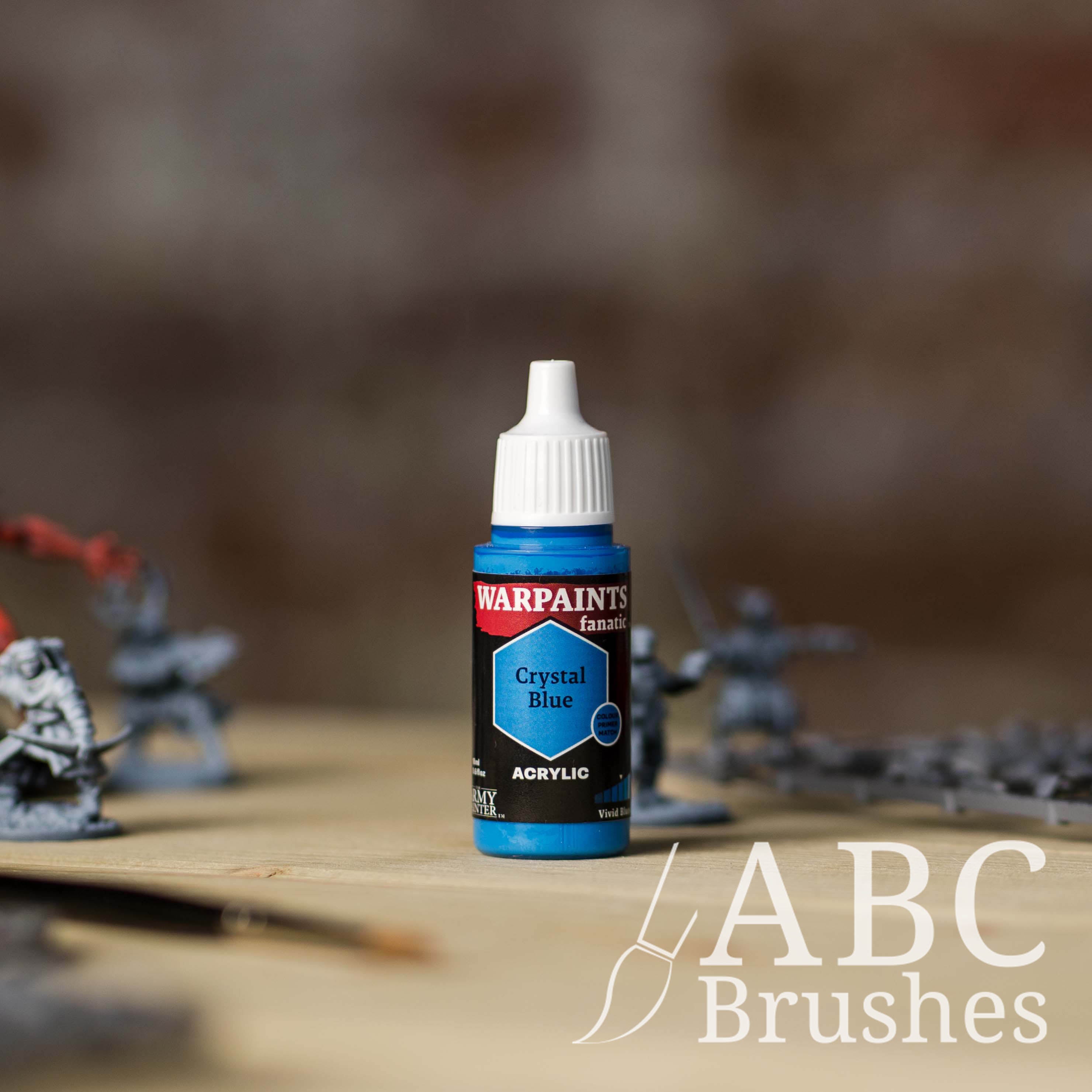 Bottle of the Army Painter Fanatics in the colour Crystal Blue with various wargaming models and an ABC paintbrush in the background, on a wooden surface.