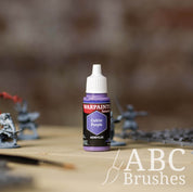 Bottle of the Army Painter Fanatics in the colour Cultist Purple with various wargaming models and an ABC paintbrush in the background, on a wooden surface.