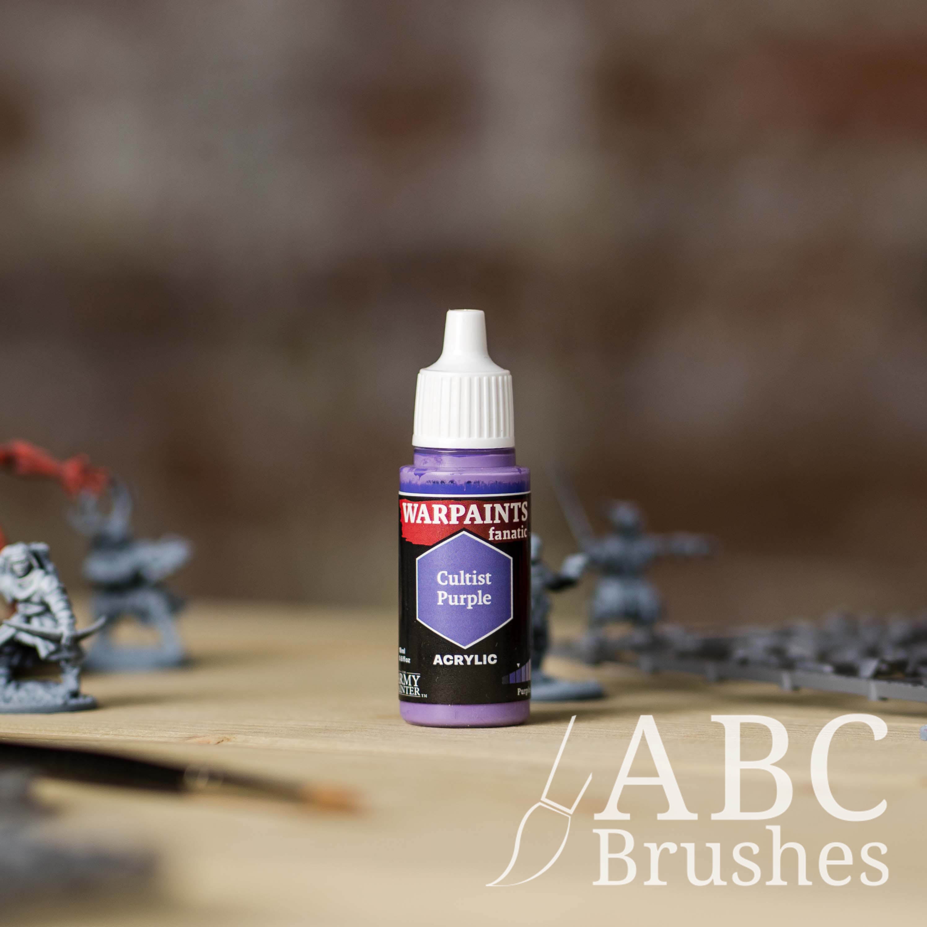 Bottle of the Army Painter Fanatics in the colour Cultist Purple with various wargaming models and an ABC paintbrush in the background, on a wooden surface.