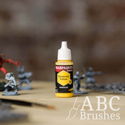 Bottle of the Army Painter Fanatics in the colour Daemonic Yellow with various wargaming models and an ABC paintbrush in the background, on a wooden surface.