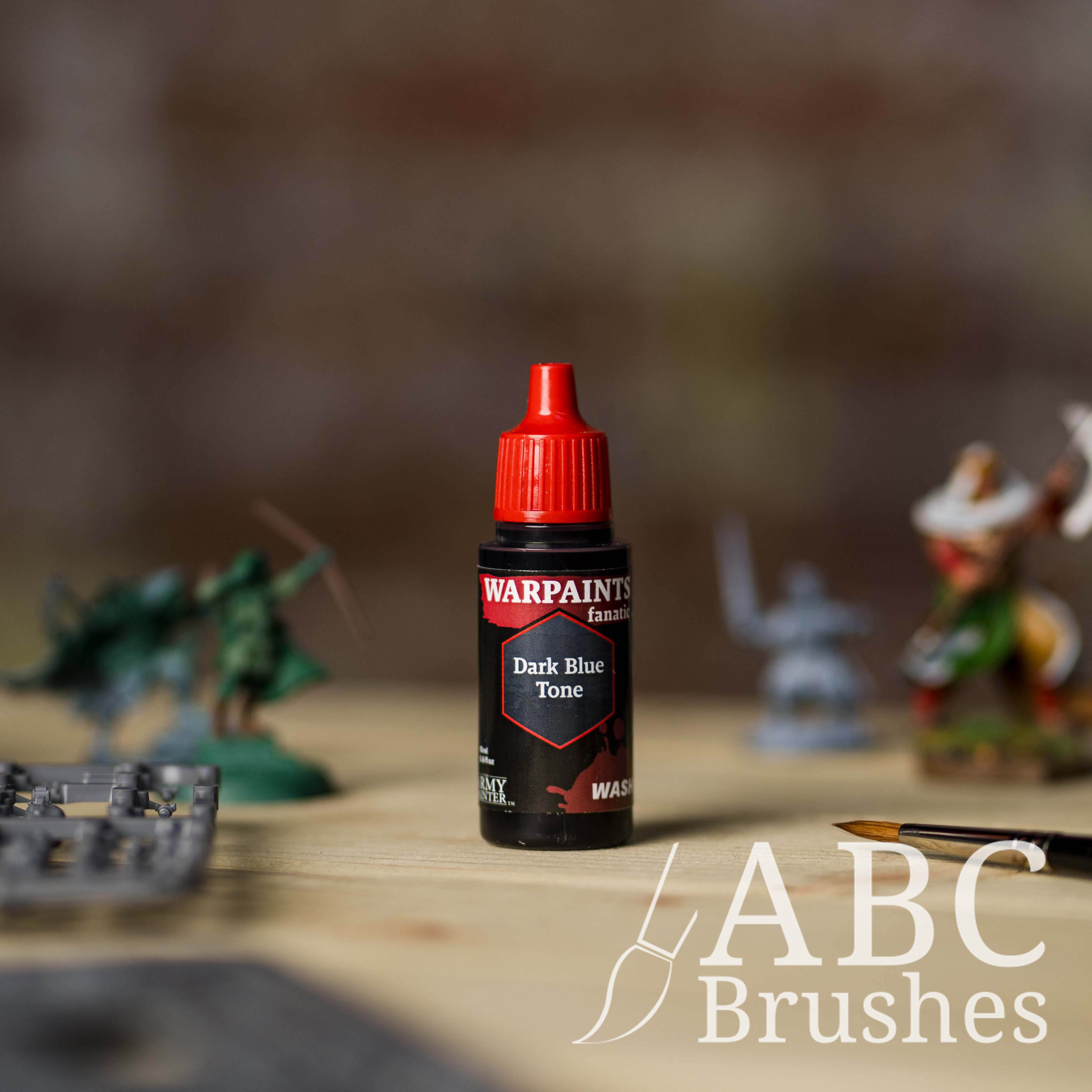 Bottle of the Army Painter Fanatics Wash in the colour Dark Blue Tone with various wargaming models and an ABC paintbrush in the background, on a wooden surface.