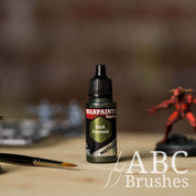 Bottle of the Army Painter Fanatics Metallic paint in the colour Dark Emerald with various wargaming models and an ABC paintbrush in the background, on a wooden surface.