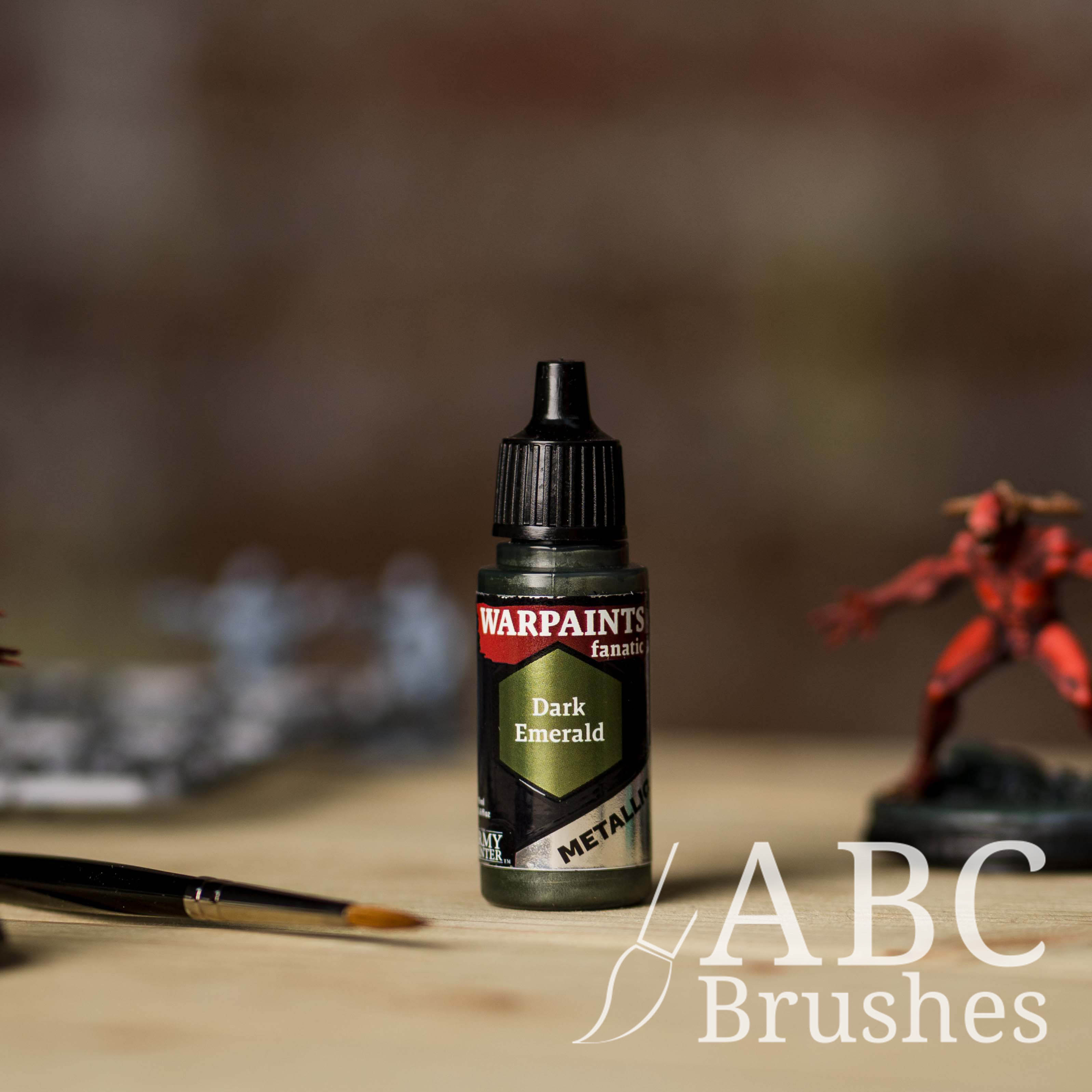 Bottle of the Army Painter Fanatics Metallic paint in the colour Dark Emerald with various wargaming models and an ABC paintbrush in the background, on a wooden surface.