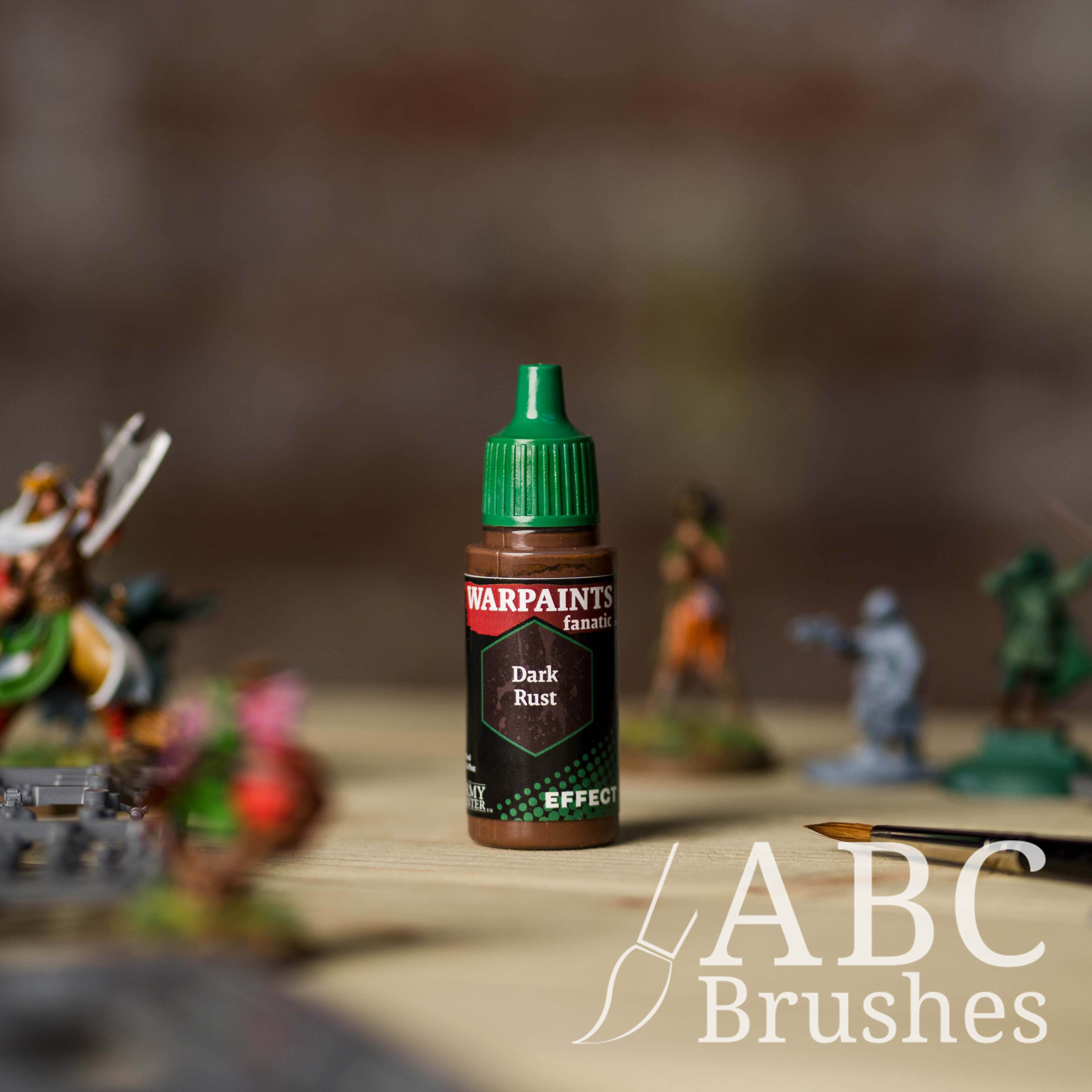 Bottle of the Army Painter Fanatics Effects in the colour Dark Rust with various wargaming models and an ABC paintbrush in the background, on a wooden surface.