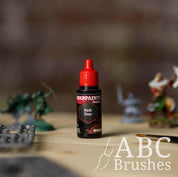 Bottle of the Army Painter Fanatics Wash in the colour Dark Tone with various wargaming models and an ABC paintbrush in the background, on a wooden surface.