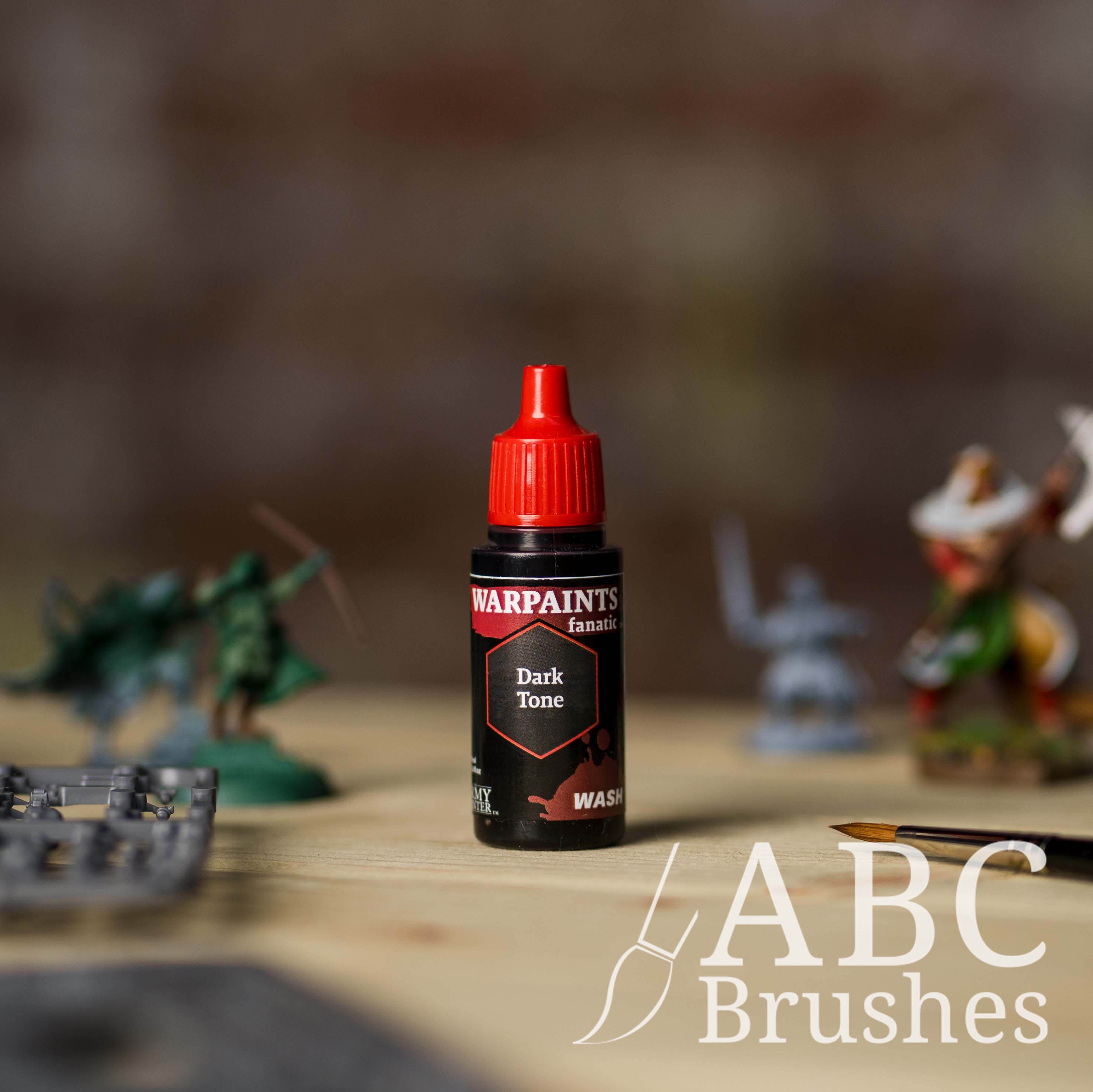 Bottle of the Army Painter Fanatics Wash in the colour Dark Tone with various wargaming models and an ABC paintbrush in the background, on a wooden surface.