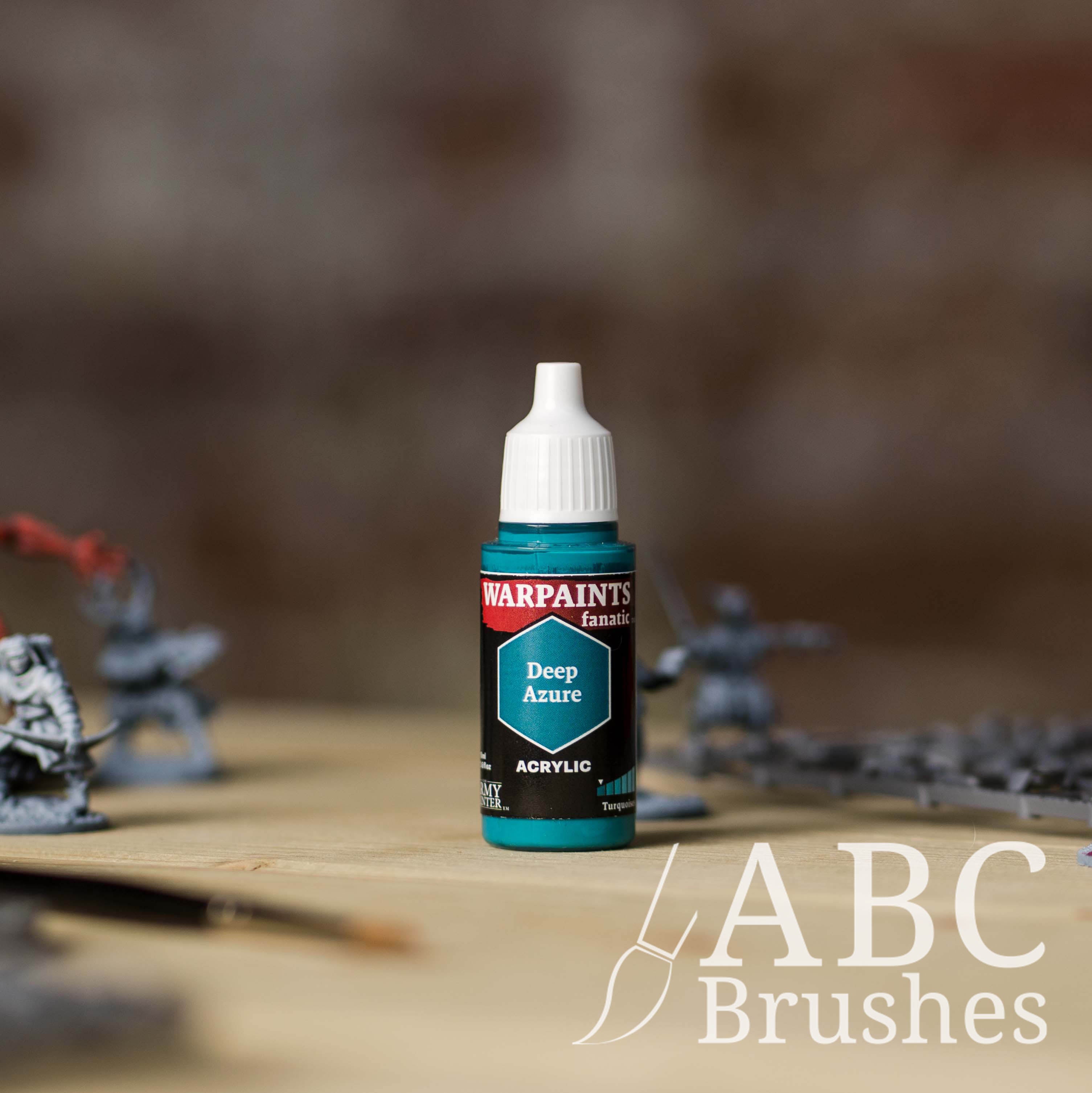 Bottle of the Army Painter Fanatics in the colour Deep Azure with various wargaming models and an ABC paintbrush in the background, on a wooden surface.