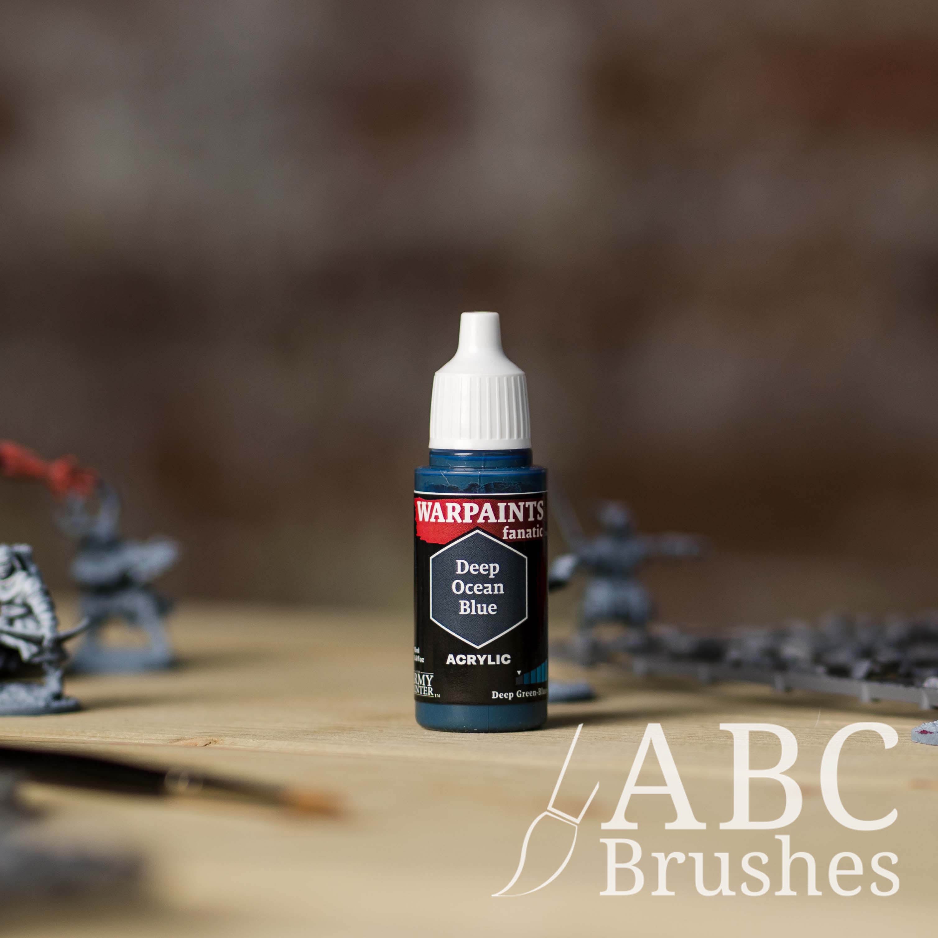 Bottle of the Army Painter Fanatics in the colour Deep Ocean Blue with various wargaming models and an ABC paintbrush in the background, on a wooden surface.