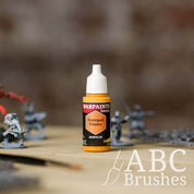 Bottle of the Army Painter Fanatics in the colour Demigod Flames with various wargaming models and an ABC paintbrush in the background, on a wooden surface.