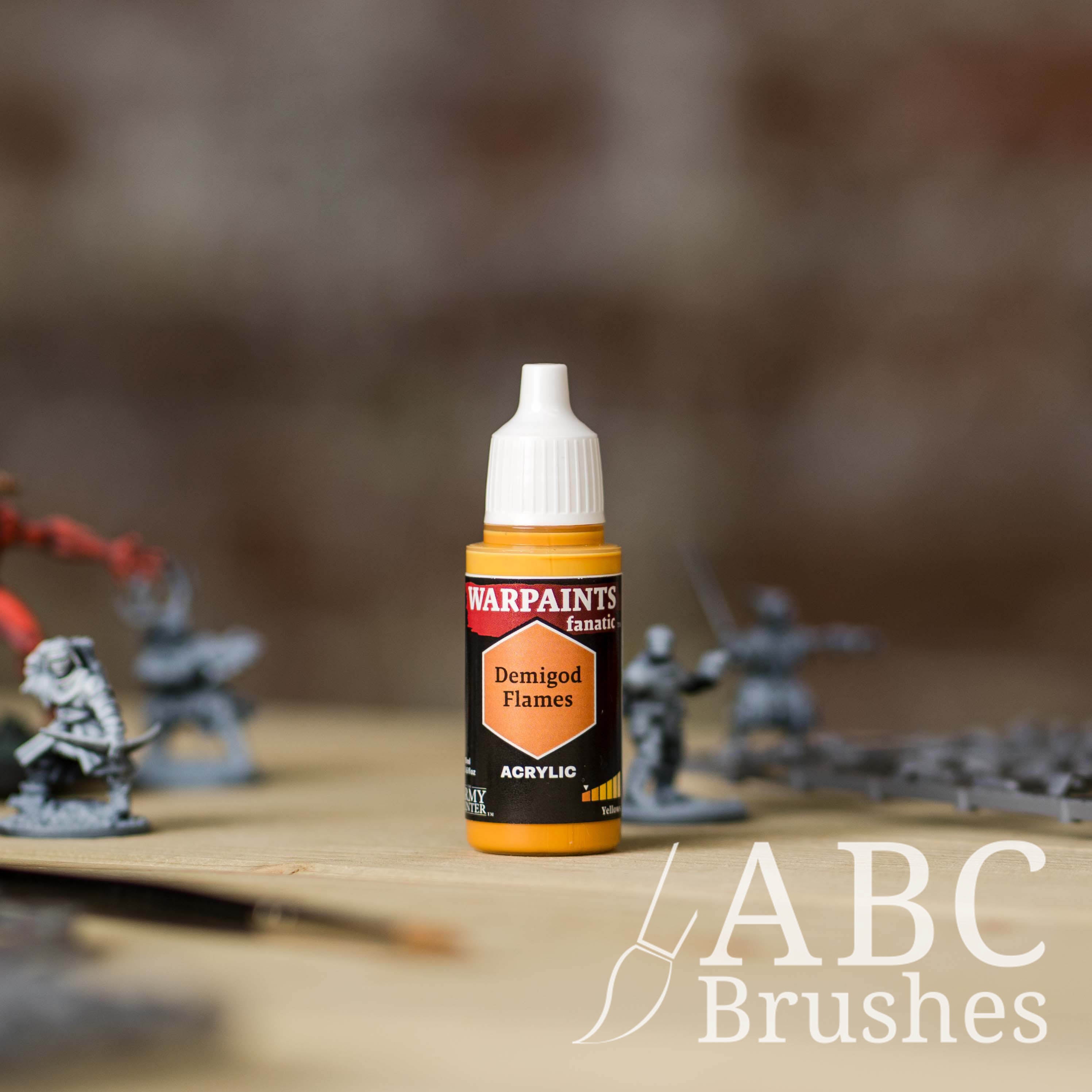 Bottle of the Army Painter Fanatics in the colour Demigod Flames with various wargaming models and an ABC paintbrush in the background, on a wooden surface.