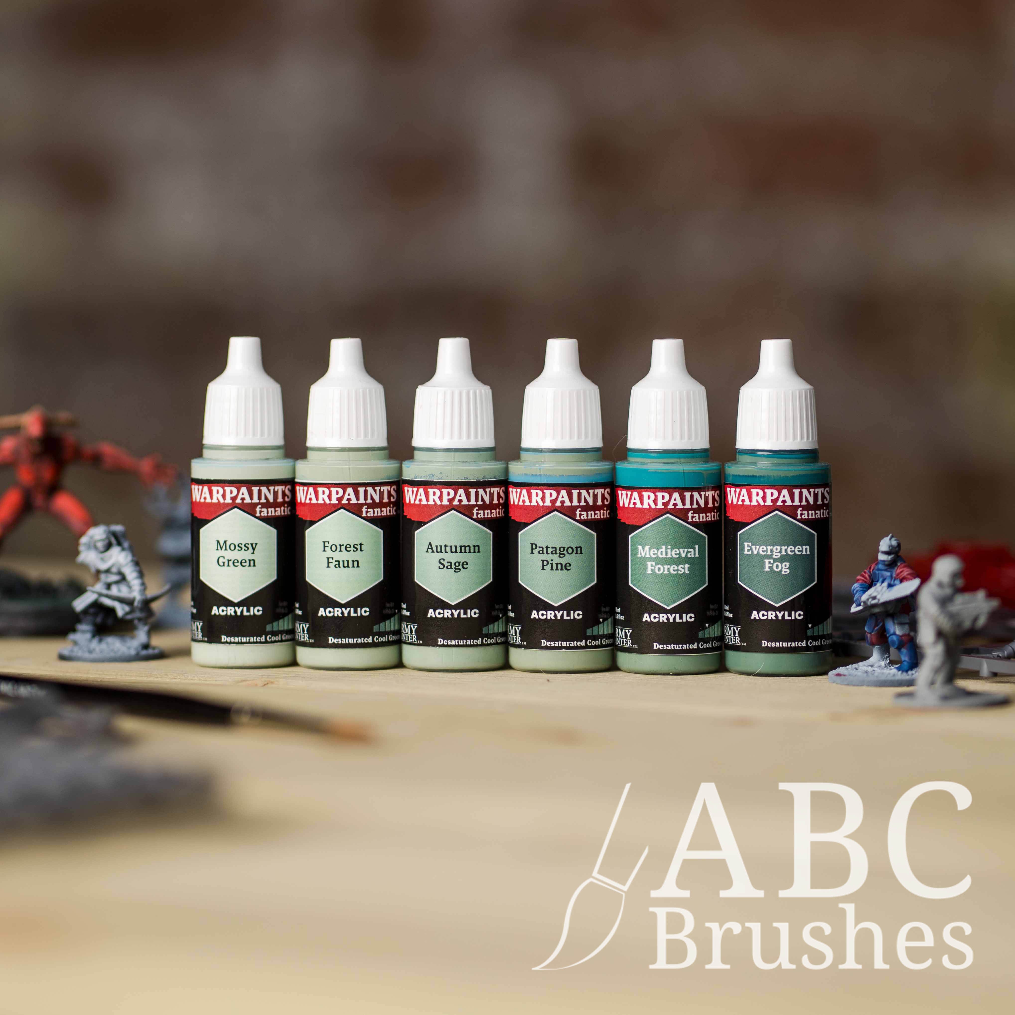 Army Painter Fanatics Cool Greens Triad on a wooden surface. There are wargaming models and an ABC paint brush next to them.