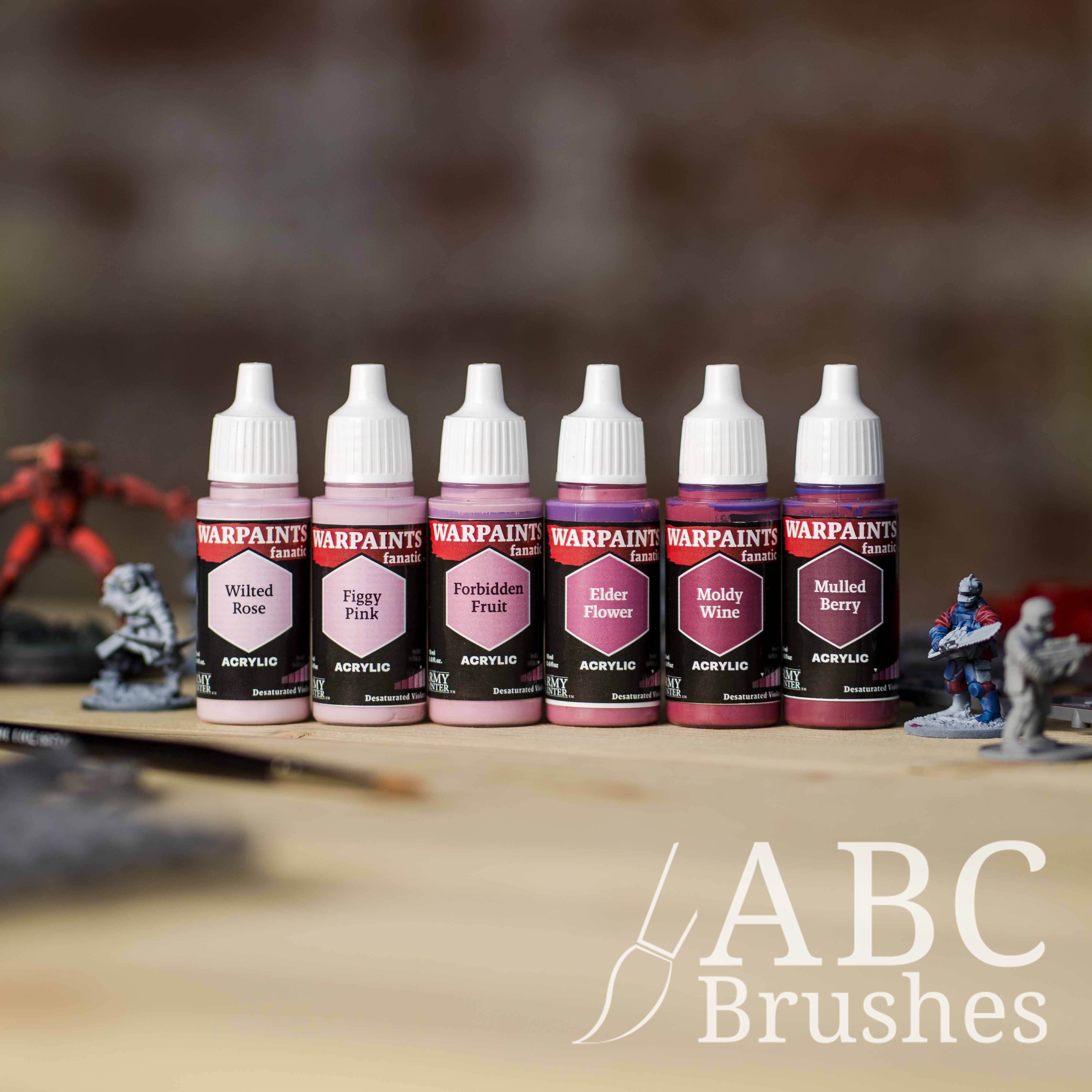 Army Painter Fanatics Desaturated Violets Triad on a wooden surface. There are wargaming models and an ABC paint brush next to them.