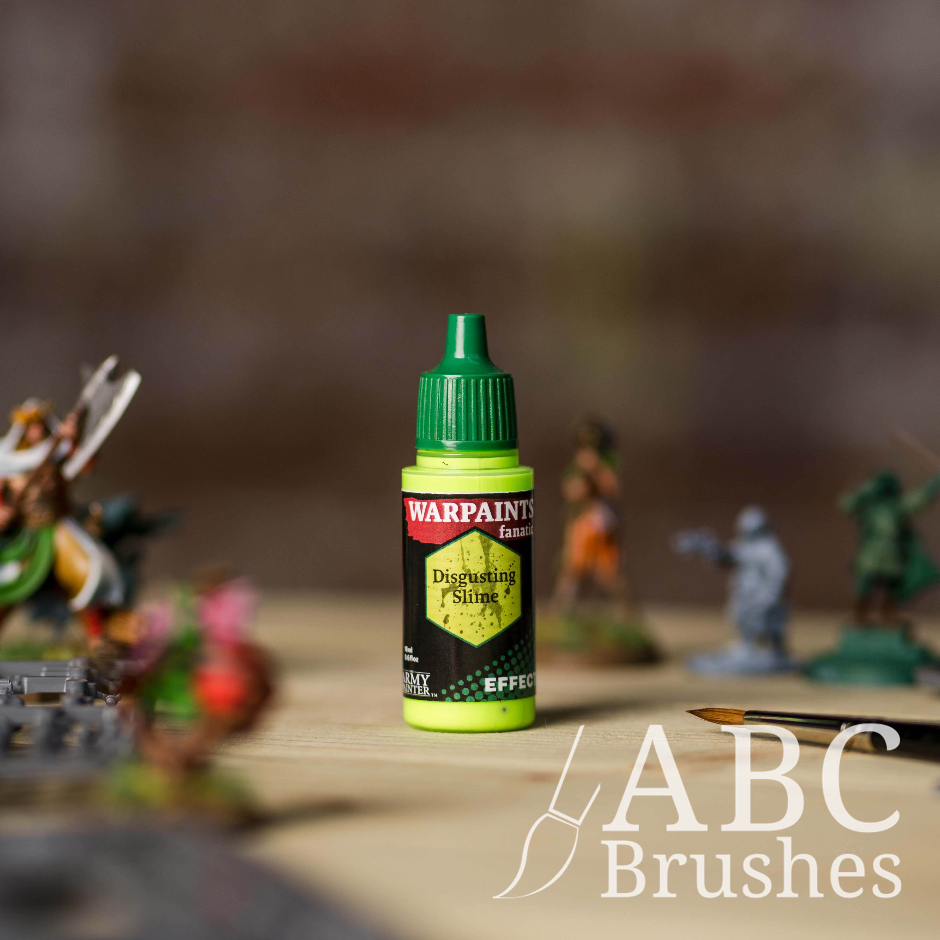 Bottle of the Army Painter Fanatics Effects in the colour Disgusting Slime with various wargaming models and an ABC paintbrush in the background, on a wooden surface.