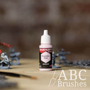 Bottle of the Army Painter Fanatics in the colour Doomfire Drab with various wargaming models and an ABC paintbrush in the background, on a wooden surface.