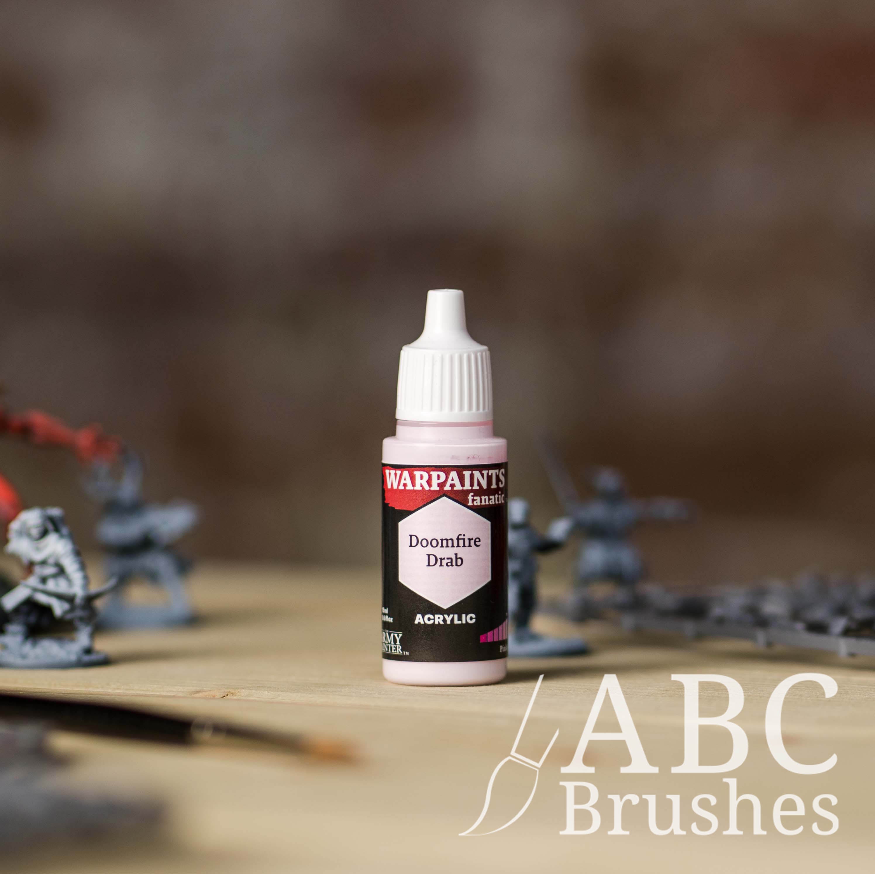 Bottle of the Army Painter Fanatics in the colour Doomfire Drab with various wargaming models and an ABC paintbrush in the background, on a wooden surface.