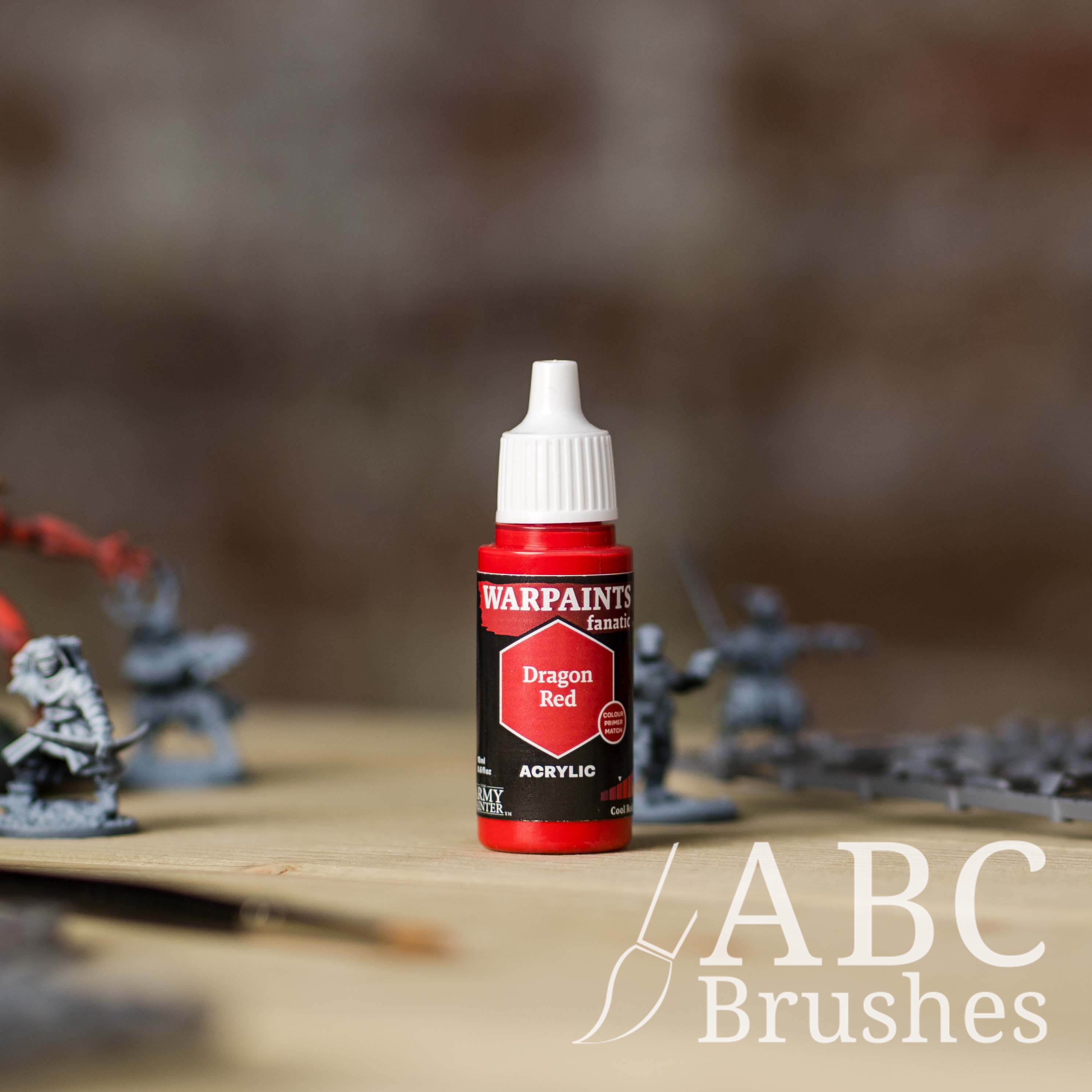 Bottle of the Army Painter Fanatics in the colour Dragon Red with various wargaming models and an ABC paintbrush in the background, on a wooden surface.