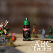 Bottle of the Army Painter Fanatics Effects in the colour Dry Blood with various wargaming models and an ABC paintbrush in the background, on a wooden surface.