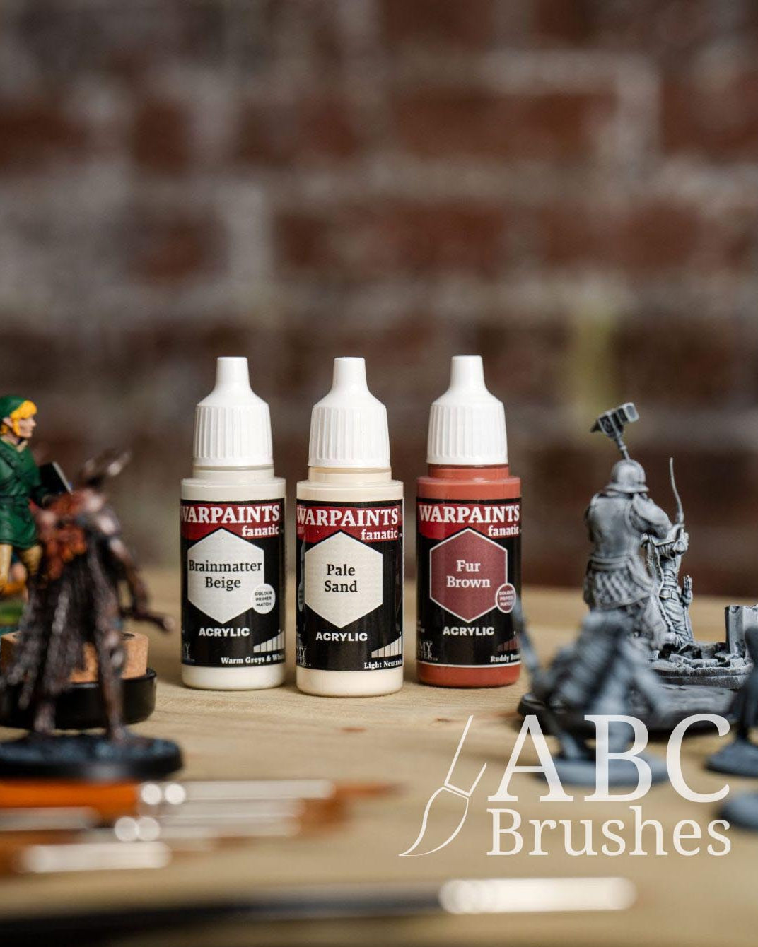 Three Warpaints paint pots in the colours, Brainmatter Beige, Pale Sand and Fur Brown on a wooden surface with miniature figures and paintbrushes in the background.