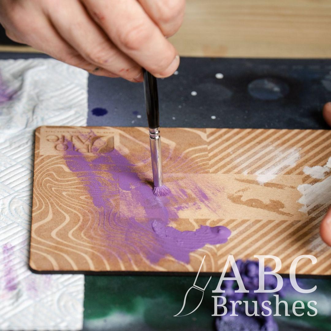Removing purple paint from a Pro Arte dry brush using a Pro Arte dry brush board, with a paper towel and purple miniature next to it. 