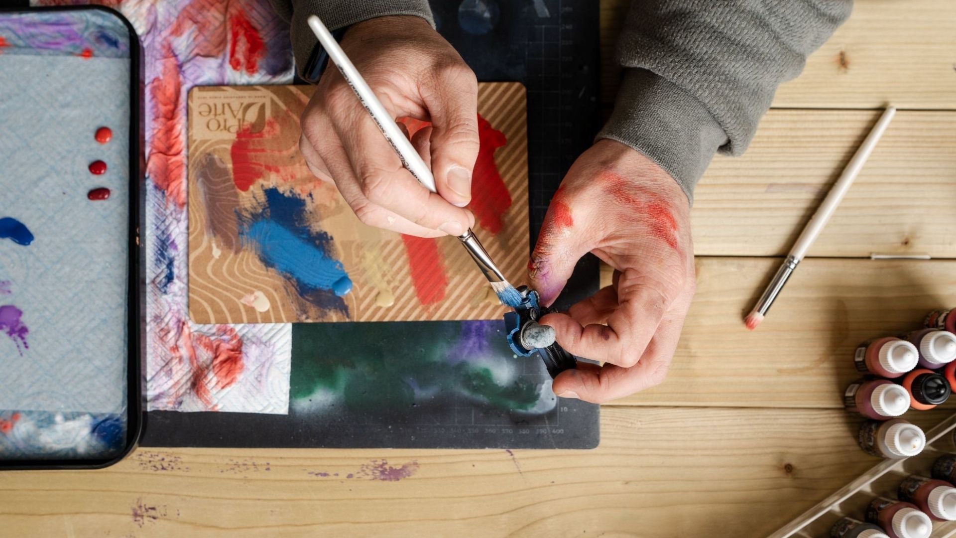 Person dry brush painting a model with a Pro Arte Masterstroke Round Foliage brush in blue paint. There is a wet palette, ABC dry brush board, selection of paints and a Pro Arte dry brush below their hands. 