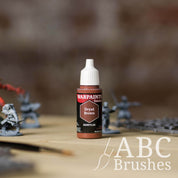 Bottle of the Army Painter Fanatics in the colour Dryad Brown with various wargaming models and an ABC paintbrush in the background, on a wooden surface.
