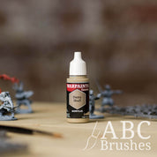 Bottle of the Army Painter Fanatics in the colour Dusty Skull with various wargaming models and an ABC paintbrush in the background, on a wooden surface.