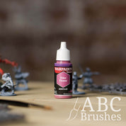 Bottle of the Army Painter Fanatics in the colour Elder Flower with various wargaming models and an ABC paintbrush in the background, on a wooden surface.