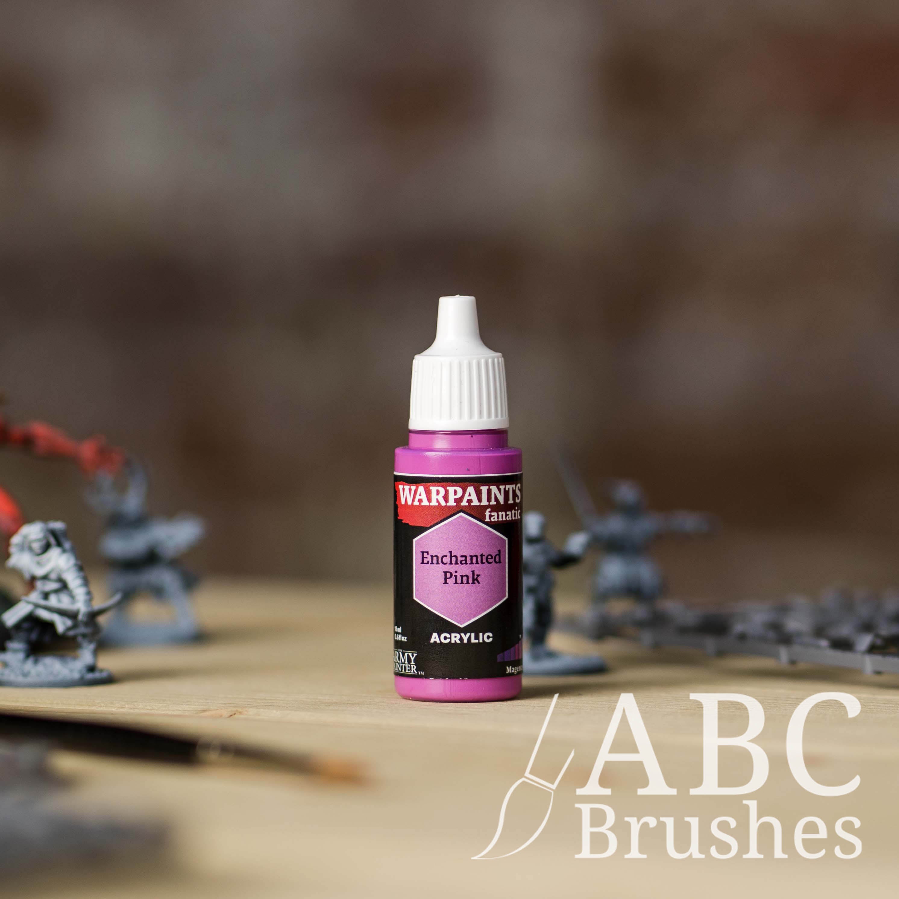 Bottle of the Army Painter Fanatics in the colour Enchanted Pink with various wargaming models and an ABC paintbrush in the background, on a wooden surface.