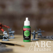 Bottle of the Army Painter Fanatics in the colour Eternal Hunt with various wargaming models and an ABC paintbrush in the background, on a wooden surface.