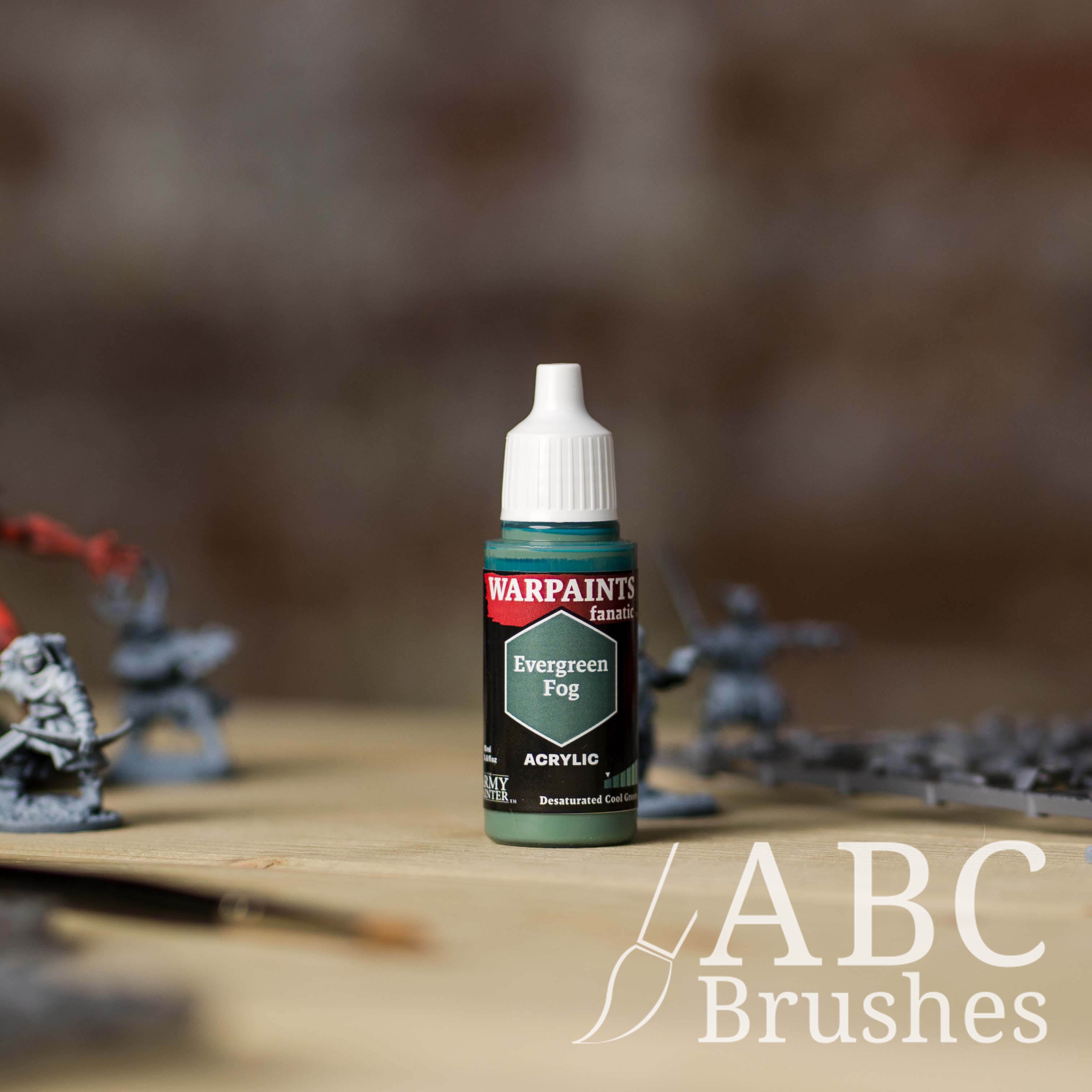 Bottle of the Army Painter Fanatics in the colour Evergreen Fog with various wargaming models and an ABC paintbrush in the background, on a wooden surface.