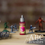 Bottle of the Army Painters Speedpaint in the colour Familiar Pink, with wargaming models and an ABC paintbrush in the background, on a wooden surface.