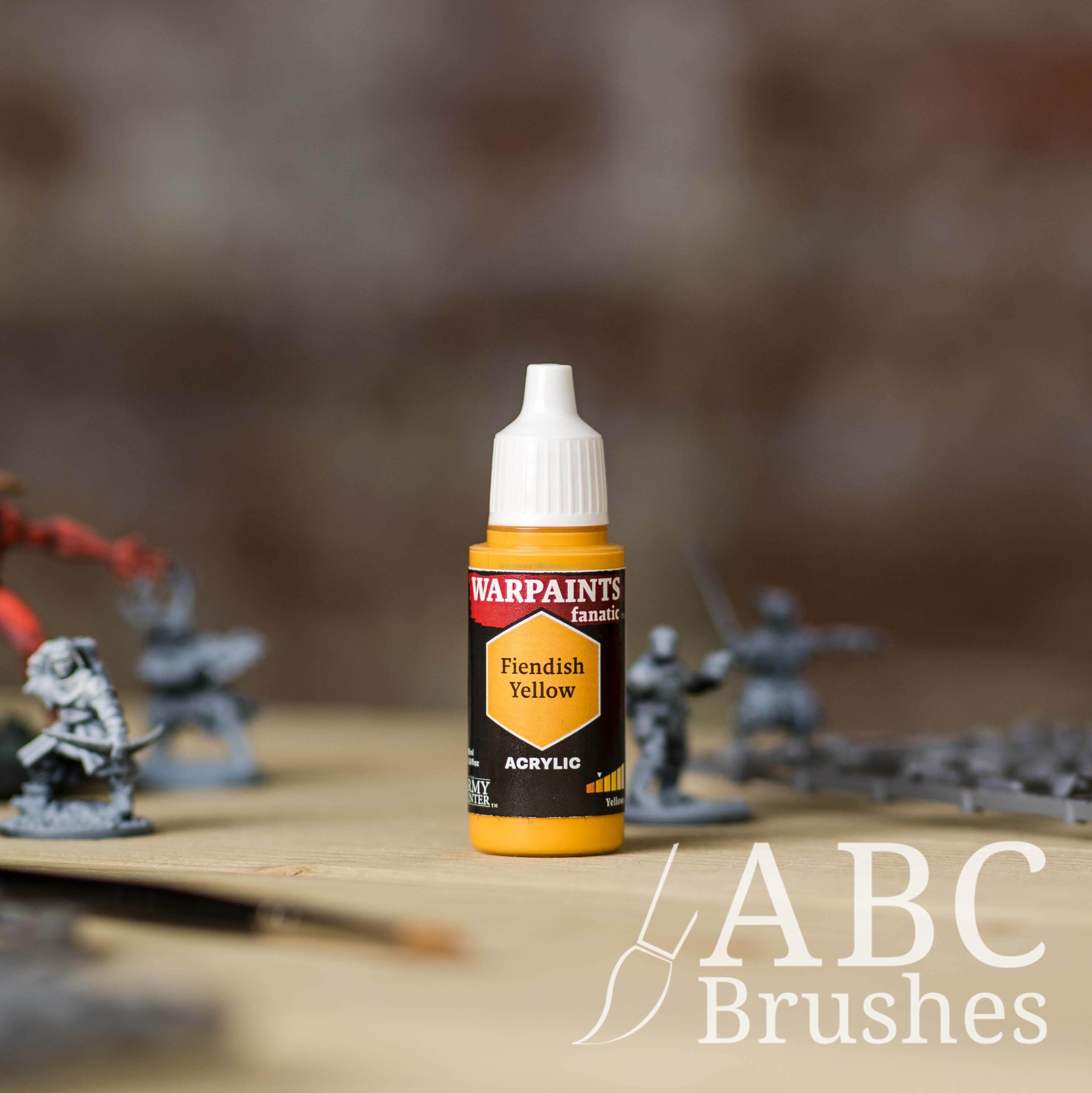 Bottle of the Army Painter Fanatics in the colour Fiendish Yellow with various wargaming models and an ABC paintbrush in the background, on a wooden surface.