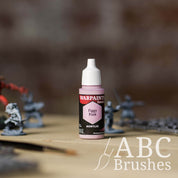 Bottle of the Army Painter Fanatics in the colour Figgy Pink with various wargaming models and an ABC paintbrush in the background, on a wooden surface.