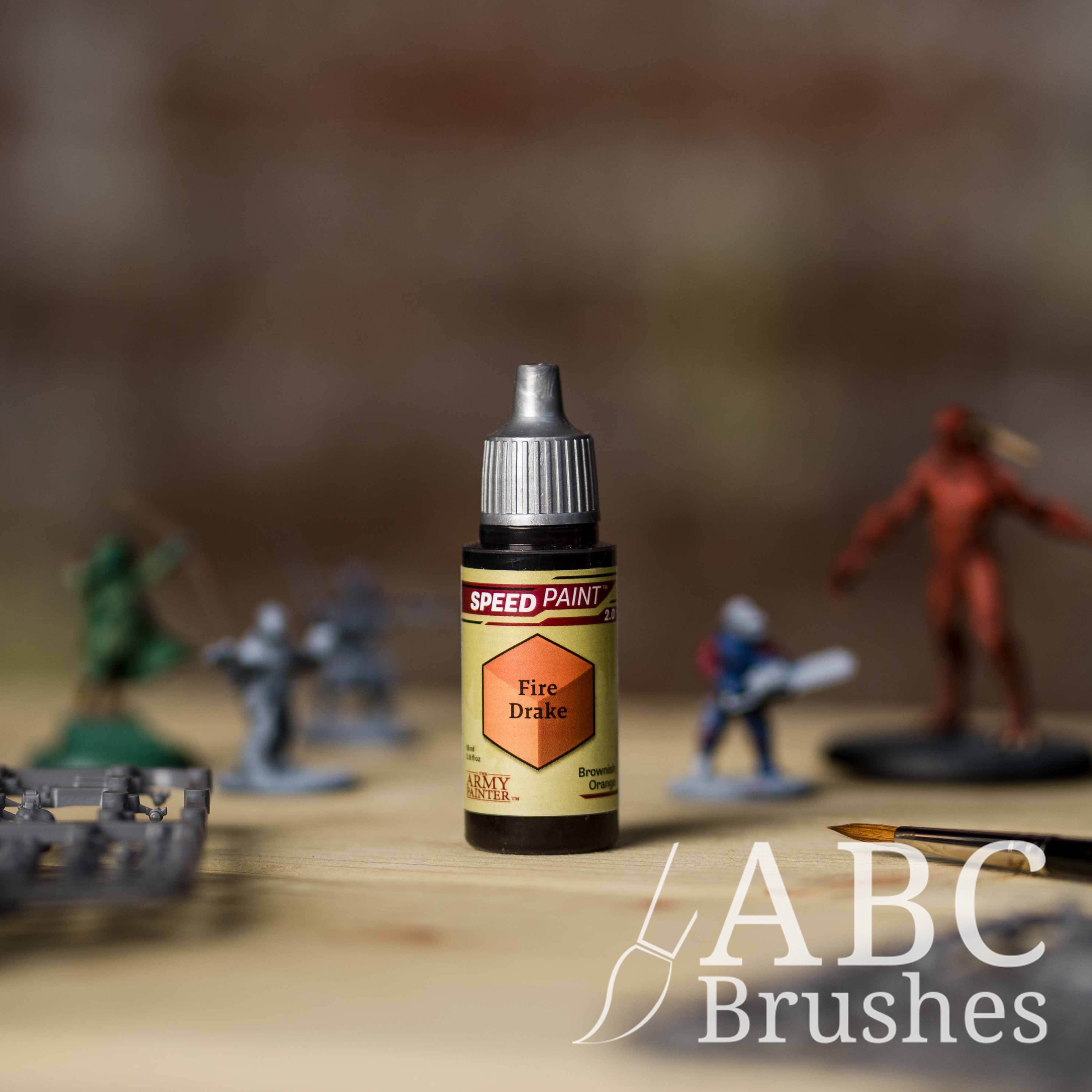 Bottle of the Army Painters Speedpaint in the colour Fire Drake, with wargaming models and an ABC paintbrush in the background, on a wooden surface.