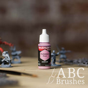 Bottle of the Army Painter Fanatics in the colour Forbidden Fruit with various wargaming models and an ABC paintbrush in the background, on a wooden surface.