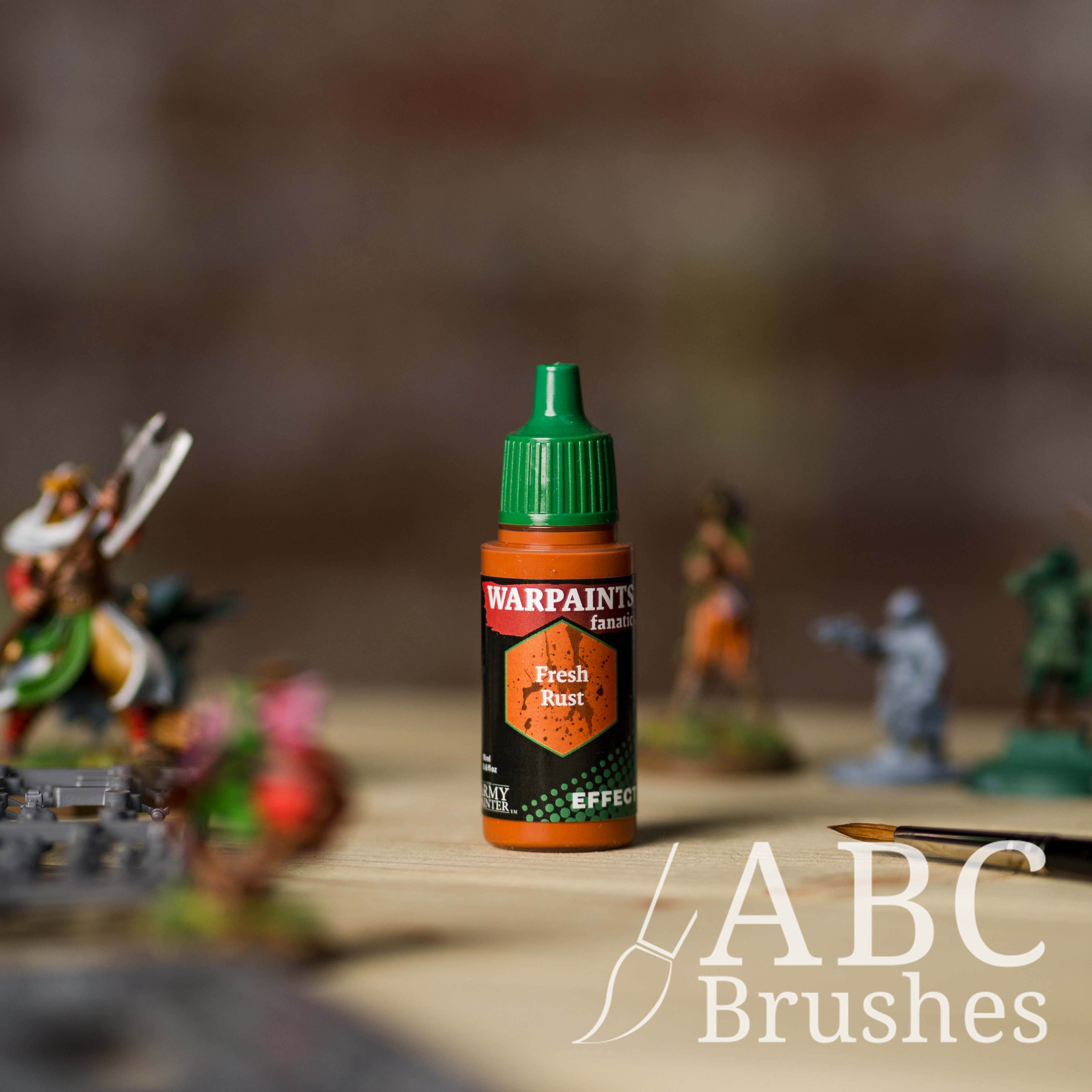 Bottle of the Army Painter Fanatics Effects in the colour Fresh Rust with various wargaming models and an ABC paintbrush in the background, on a wooden surface.