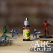 Bottle of the Army Painters Speedpaint in the colour Ghillie Dew, with wargaming models and an ABC paintbrush in the background, on a wooden surface.