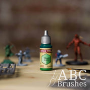 Bottle of the Army Painters Speedpaint in the colour Ghoul Green, with wargaming models and an ABC paintbrush in the background, on a wooden surface.