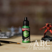 Bottle of the Army Painter Fanatics Metallic paint in the colour Glittering Green with various wargaming models and an ABC paintbrush in the background, on a wooden surface.