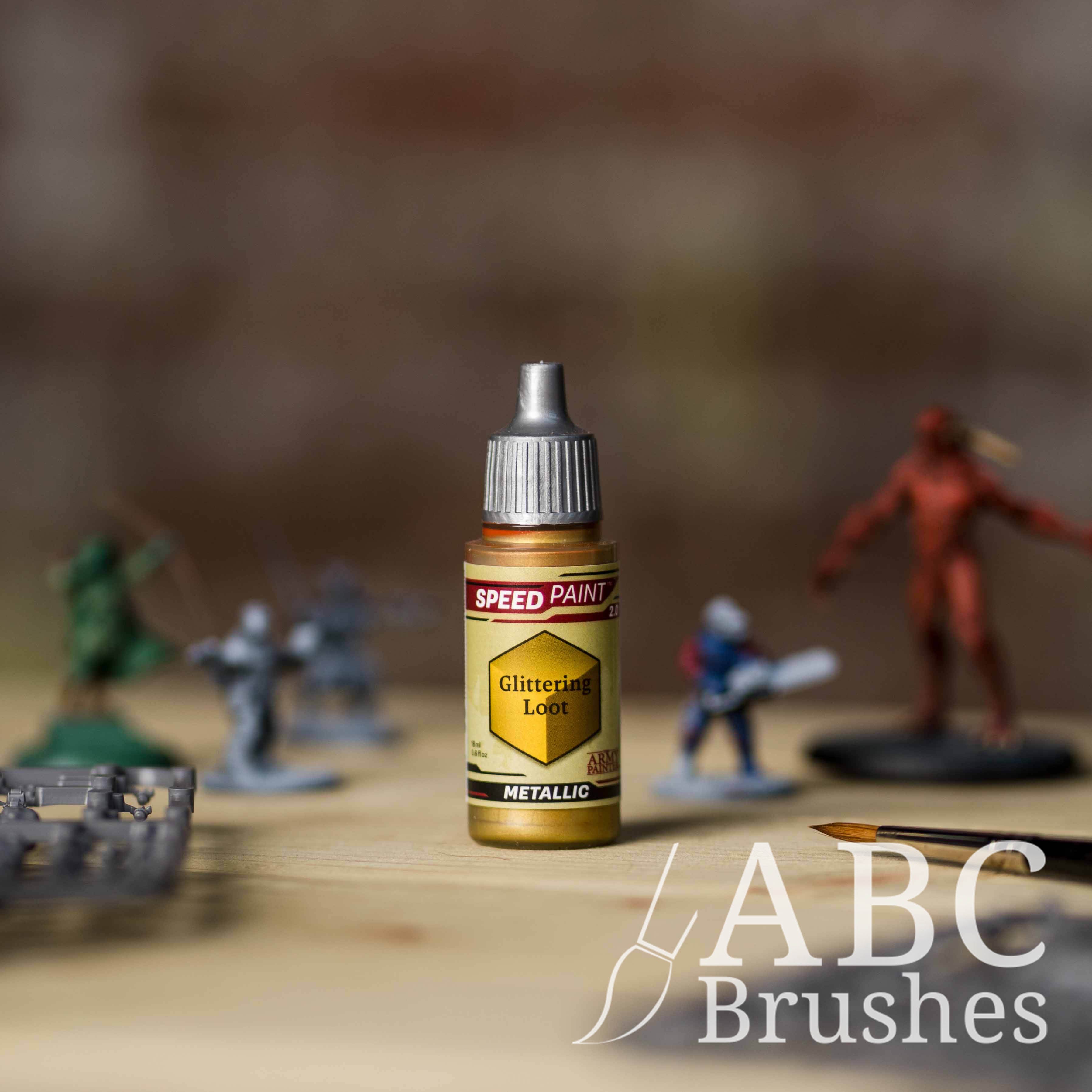 Bottle of the Army Painters Metallic Speedpaint in the colour Glittering Loot, with wargaming models and an ABC paintbrush in the background, on a wooden surface.