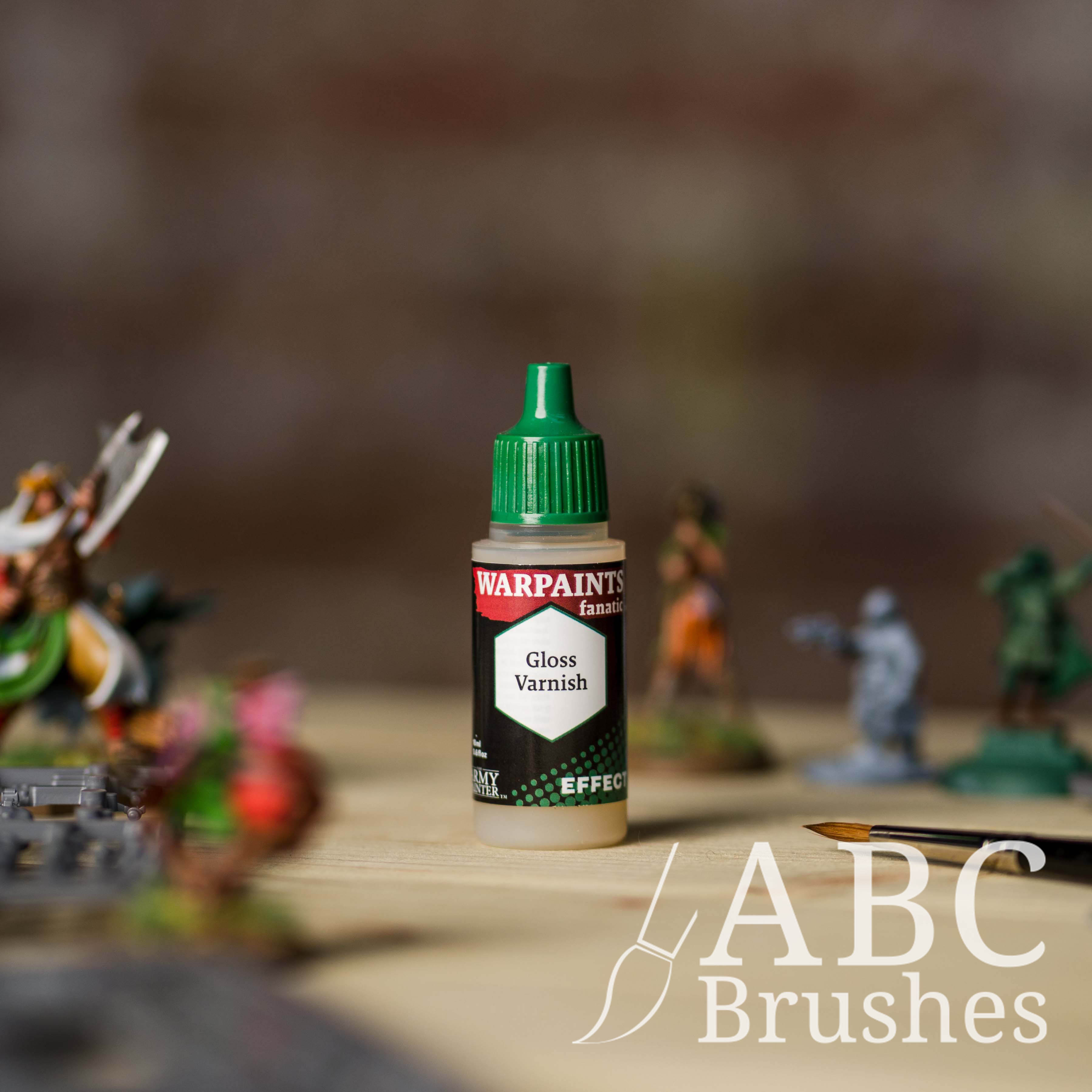 Bottle of the Army Painter Fanatics Effects Gloss Varnish with various wargaming models and an ABC paintbrush in the background, on a wooden surface.