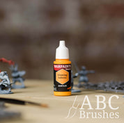 Bottle of the Army Painter Fanatics in the colour Glowing Inferno with various wargaming models and an ABC paintbrush in the background, on a wooden surface.