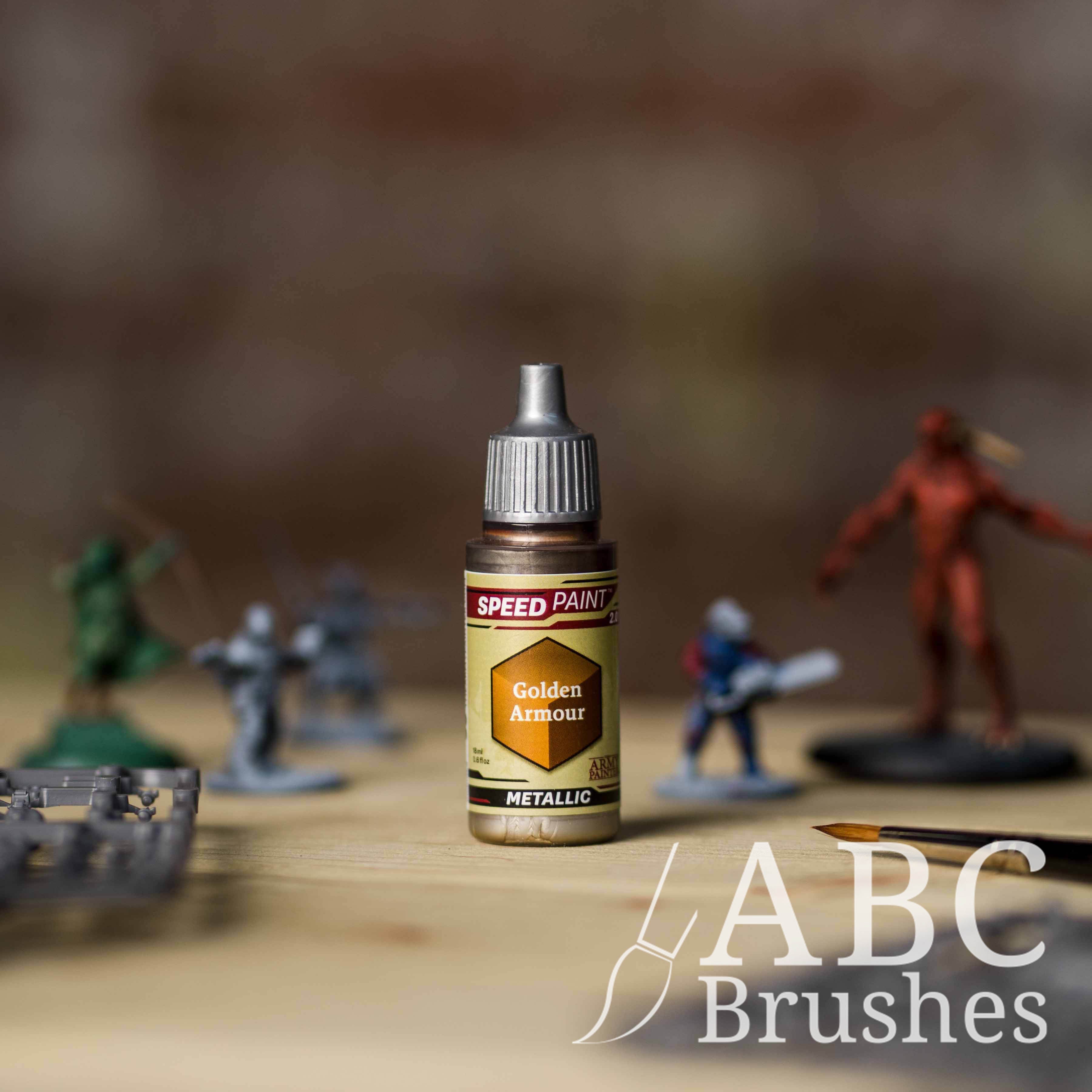 Bottle of the Army Painters Metallic Speedpaint in the colour Golden Armour, with wargaming models and an ABC paintbrush in the background, on a wooden surface.