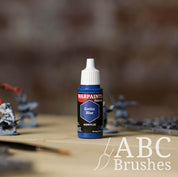 Bottle of the Army Painter Fanatics in the colour Gothic Blue with various wargaming models and an ABC paintbrush in the background, on a wooden surface.