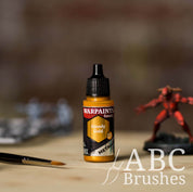 Bottle of the Army Painter Fanatics Metallic paint in the colour Greedy Gold with various wargaming models and an ABC paintbrush in the background, on a wooden surface.