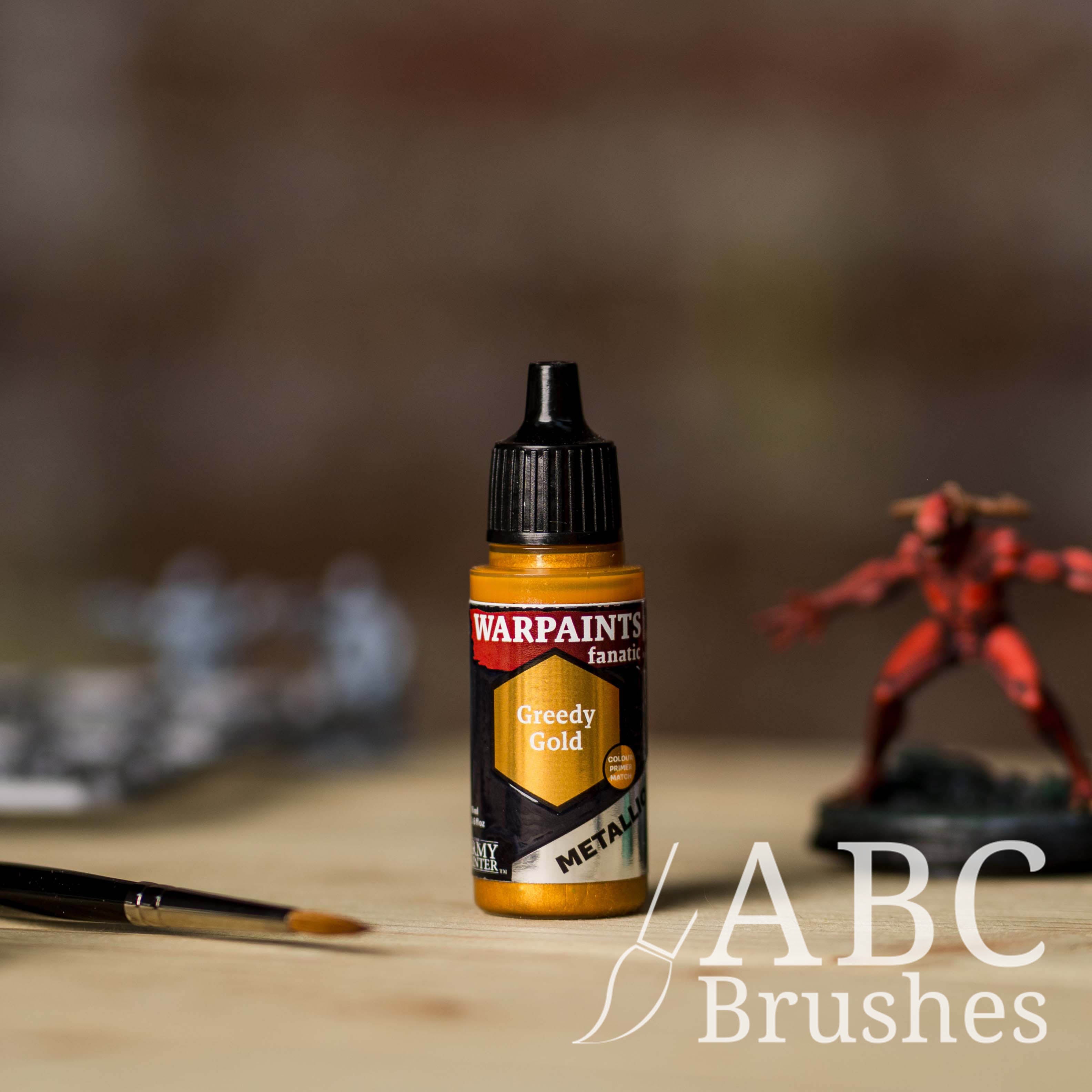 Bottle of the Army Painter Fanatics Metallic paint in the colour Greedy Gold with various wargaming models and an ABC paintbrush in the background, on a wooden surface.