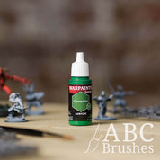 Bottle of the Army Painter Fanatics in the colour Greenskin with various wargaming models and an ABC paintbrush in the background, on a wooden surface.