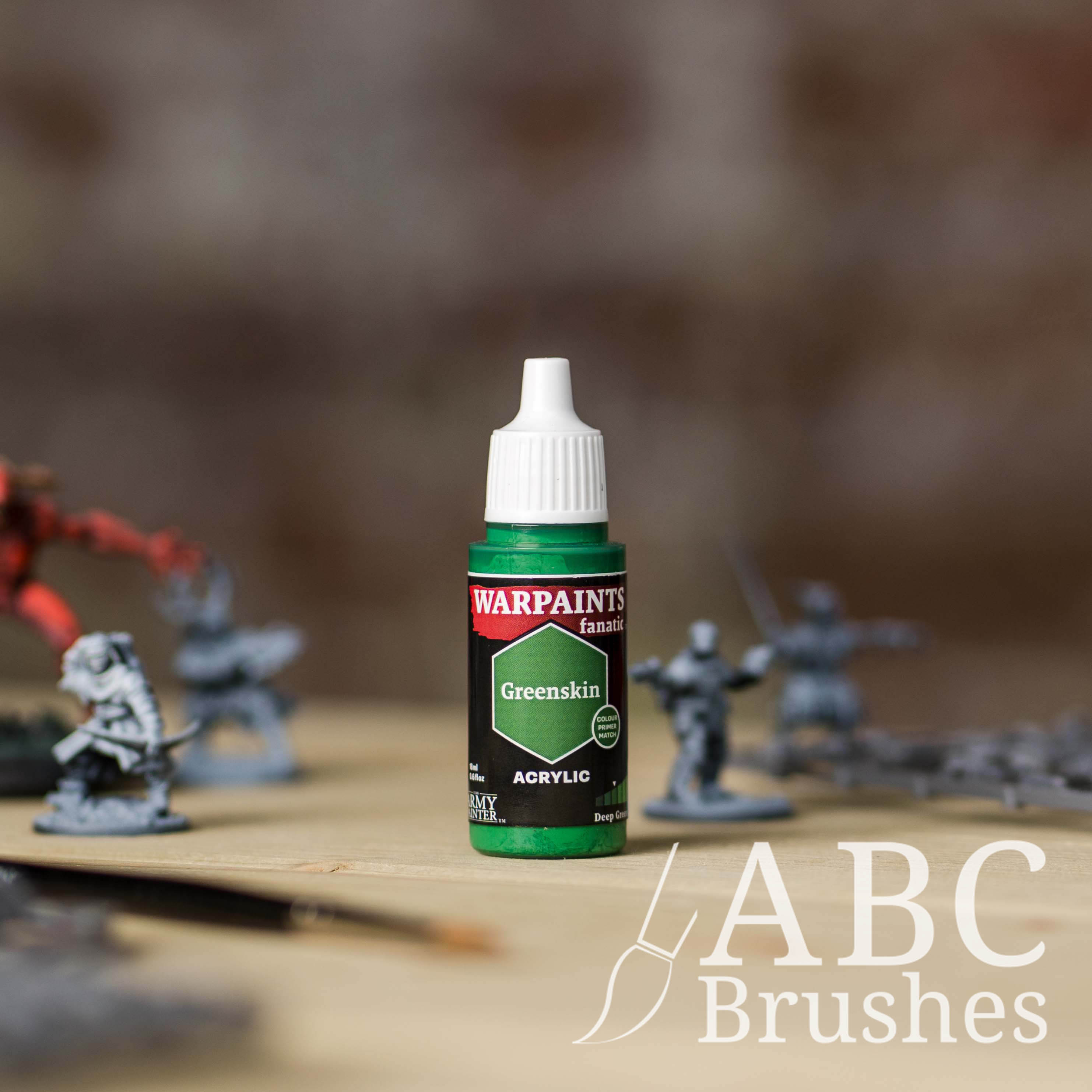 Bottle of the Army Painter Fanatics in the colour Greenskin with various wargaming models and an ABC paintbrush in the background, on a wooden surface.