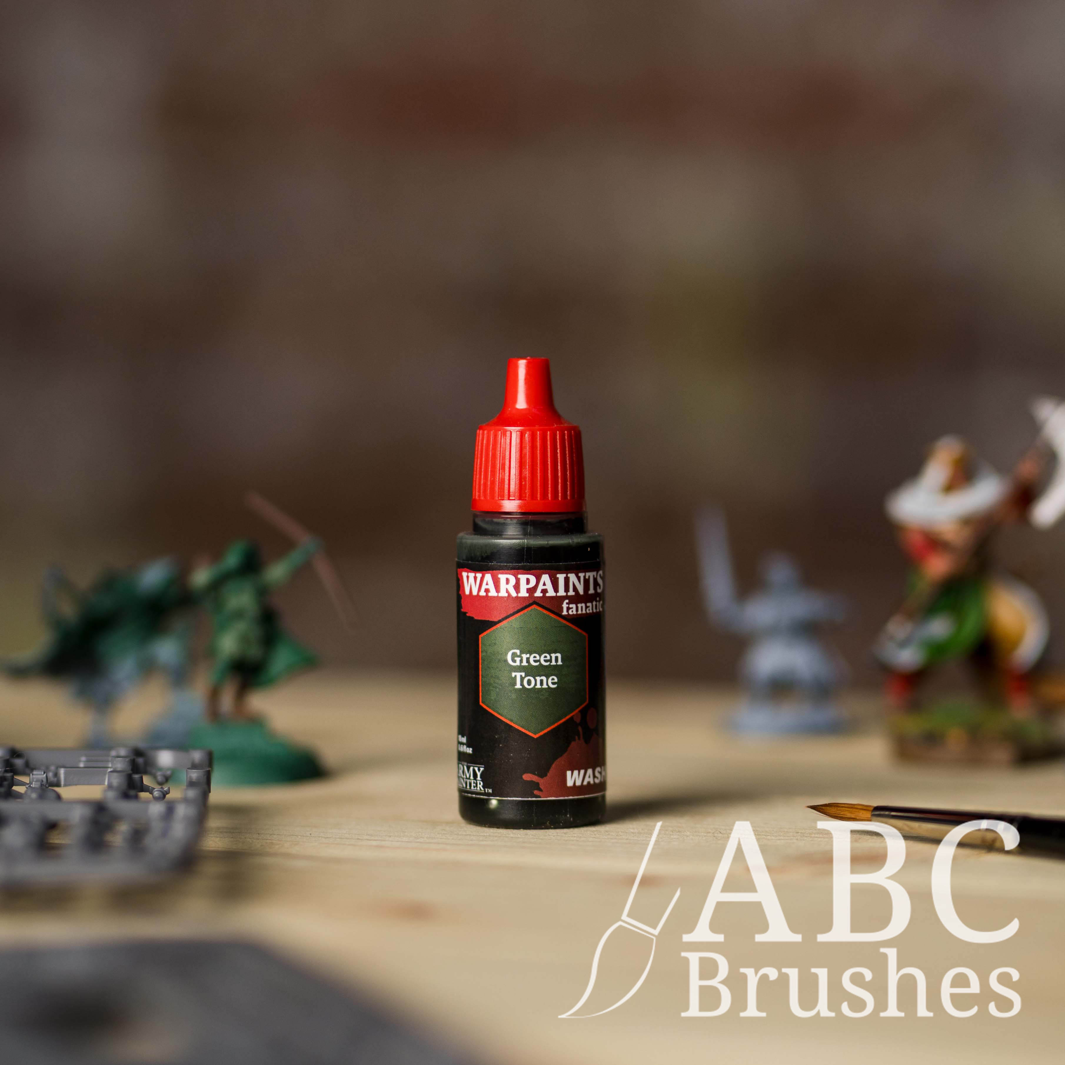 Bottle of the Army Painter Fanatics Wash in the colour Green Tone with various wargaming models and an ABC paintbrush in the background, on a wooden surface.